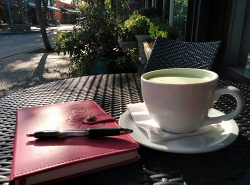 A mug of matcha green tea on a plate with a red notebook and pen on an outdoor table.